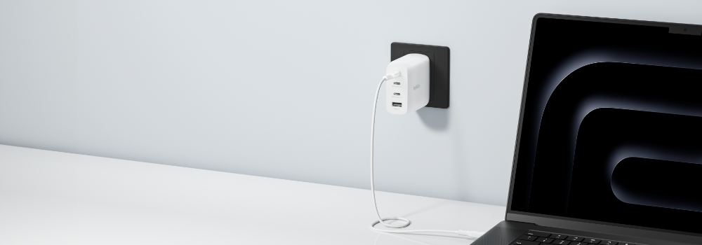 White Belkin BoostCharge Pro multi-port USB-C wall charger plugged into an outlet on a clean white wall, with a USB-C cable running to a slim laptop on a desk. Minimalist studio aesthetic.