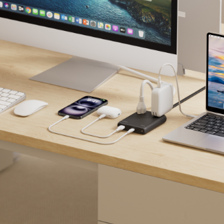 A tidy desk setup featuring a desktop computer, laptop, keyboard, mouse, and smartphone, all powered through a compact multi-port charging hub placed between the devices. Several cables connect the hub to the laptop, phone, and other electronics on the light wood surface.