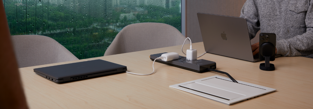 A modern conference room setting with a wooden table and two gray chairs. A person is working on a MacBook connected to a slim black power strip with multiple devices plugged in, including phone chargers. A Dell laptop is closed on the table, and a mounted phone stand with a smartphone is placed nearby. The room has large windows with raindrops visible on the glass, suggesting a rainy day outside.