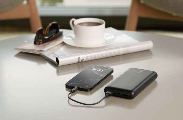 A sleek black smartphone is charging via a cable connected to a slim black Belkin power bank on a modern gray table. Next to them is a rolled-up magazine, a white cup and saucer filled with coffee, and a pair of sunglasses resting on top of another magazine. The background suggests a cozy indoor setting with soft seating and natural light.