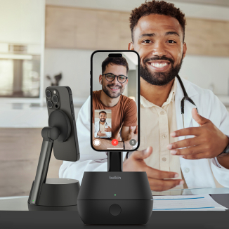 The image depicts a video call setup using a smartphone mounted on an auto-tracking stand. A smiling doctor wearing a white coat is in the background, gesturing as he engages in a virtual consultation. The smartphone screen shows a person on the other end of the video call. The sleek, black auto-tracking stand prominently displays the Belkin logo, emphasizing hands-free convenience in a professional environment.