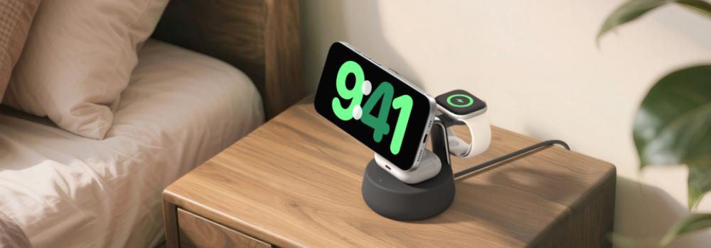 A black Belkin 3-in-1 wireless charging stand on a wooden nightstand, holding a smartphone in landscape mode displaying the time 9:41 and charging an Apple Watch beside it, next to a neatly made bed in a cozy bedroom setting.