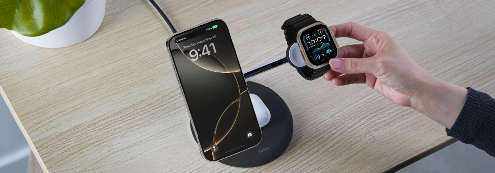 A person places an Apple Watch onto a Belkin 3-in-1 wireless charging stand on a wooden desk. The stand is already charging an iPhone and a pair of AirPods. A potted plant is partially visible in the background.