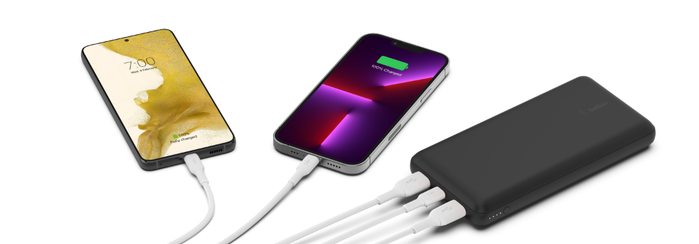 A black Belkin power bank charging two smartphones simultaneously via multiple USB cables. One phone displays a green battery icon indicating full charge, while the other shows a charging status. The power bank features multiple ports, highlighting its ability to charge multiple devices at once. The background is clean and minimalistic.