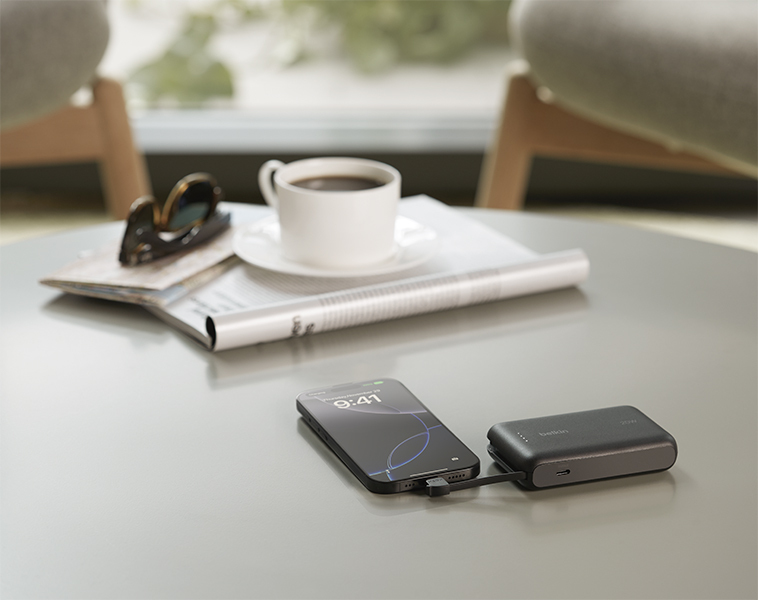 A smartphone is charging on a table via a black Belkin portable power bank. Next to it are a rolled-up magazine, a white coffee cup and saucer, and a pair of sunglasses. The setting appears to be a cozy, modern lounge or cafe with soft seating in the background.