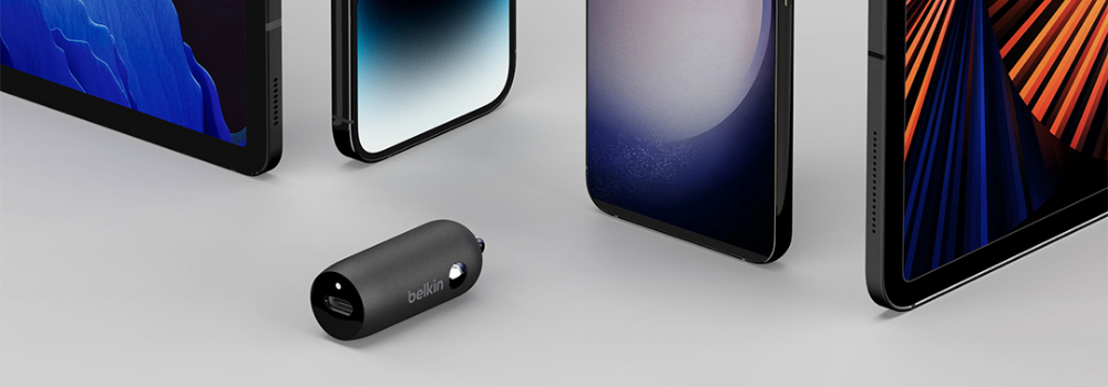 A Belkin USB car charger is displayed on a flat surface, surrounded by various mobile devices including tablets and smartphones, highlighting its compatibility with multiple device types.