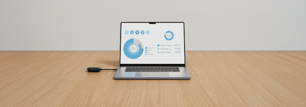 A laptop on a wooden desk connected to a small external device via cable, displaying a data dashboard on the screen.