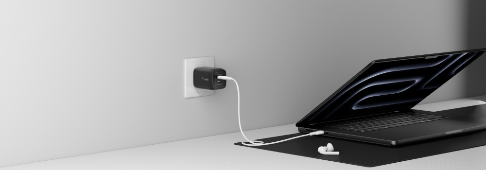 A Belkin dual USB-C wall charger plugged into a wall outlet, charging a black laptop via a white USB-C cable. The laptop is partially open and resting on a desk with a black desk mat and a pair of white wireless earbuds nearby.