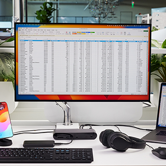A modern office desk setup featuring a large external monitor displaying an Excel spreadsheet, a MacBook laptop also showing a spreadsheet, and various accessories. The workspace includes a Belkin docking station, a wireless keyboard, a mouse, a pair of headphones, and a smartphone on a charging stand. The environment is well-lit, with green plants in the background, creating a clean and organized workspace.