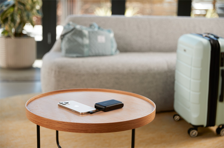 A smartphone connected to a compact Belkin power bank on a round wooden table, with travel bags and a suitcase in the background, suggesting a travel-friendly charging setup.