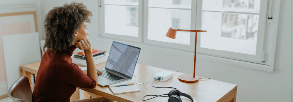 The image depicts a woman sitting at a wooden desk in a bright, airy room. She is using a laptop while resting her chin on her hand, seemingly focused on her work. The desk is organized, with a small charging hub connected to her laptop, a pair of headphones, and a notebook nearby. A modern, orange desk lamp adds a pop of color to the setup. Large windows in the background let in plenty of natural light, creating a calm and productive atmosphere.