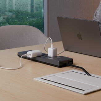 A Belkin black surge protector on a wooden desk with multiple devices plugged in, including chargers and a MacBook, near a window with raindrops visible on the glass.