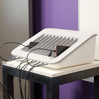 A white multi-device charging station with vertical slots, holding several smartphones connected with charging cables, placed on a small table against a purple and white wall.