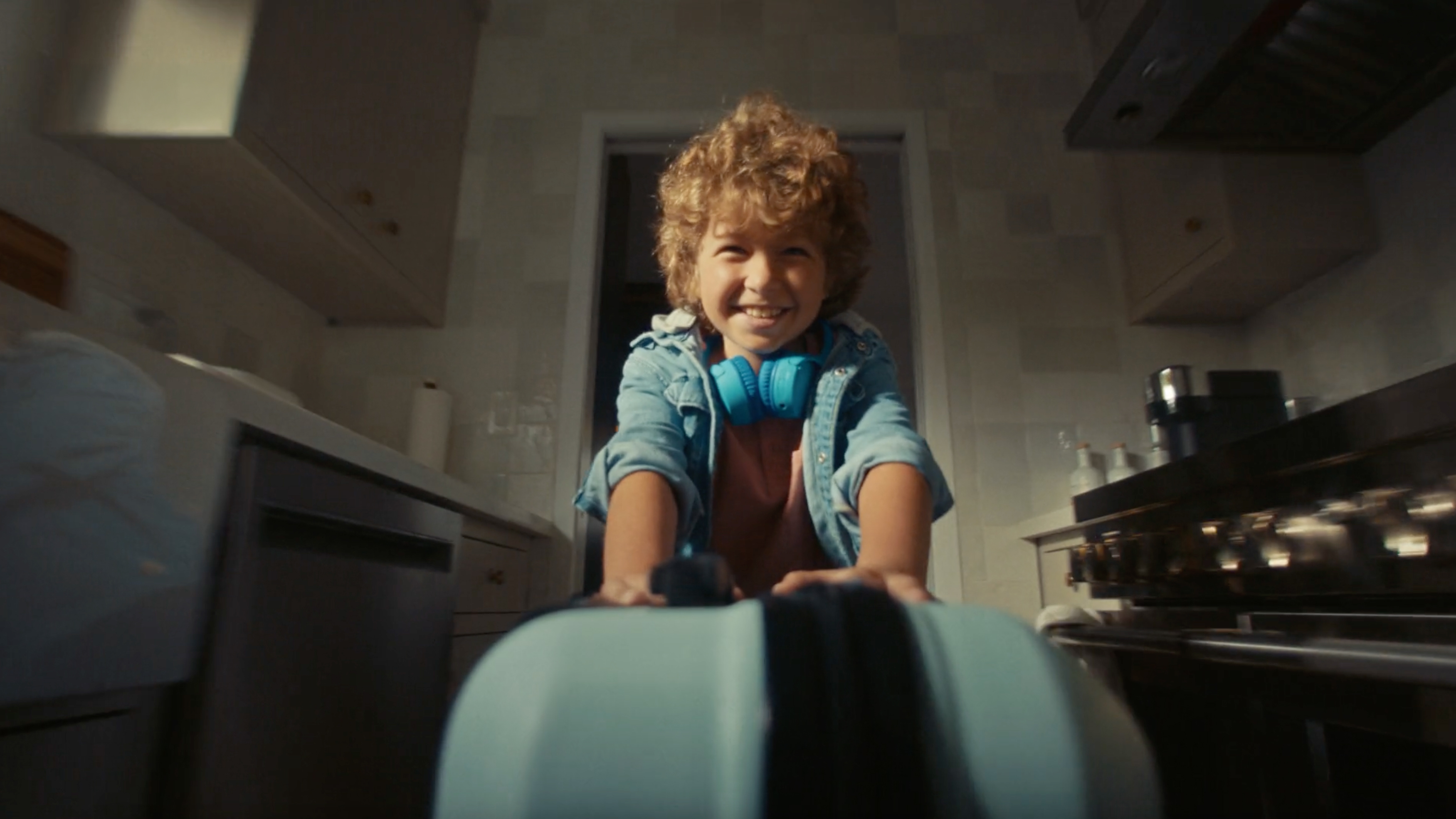 Young boy with curly hair smiling and pushing a blue suitcase toward the camera in a kitchen, wearing a denim jacket and bright blue headphones around his neck.
