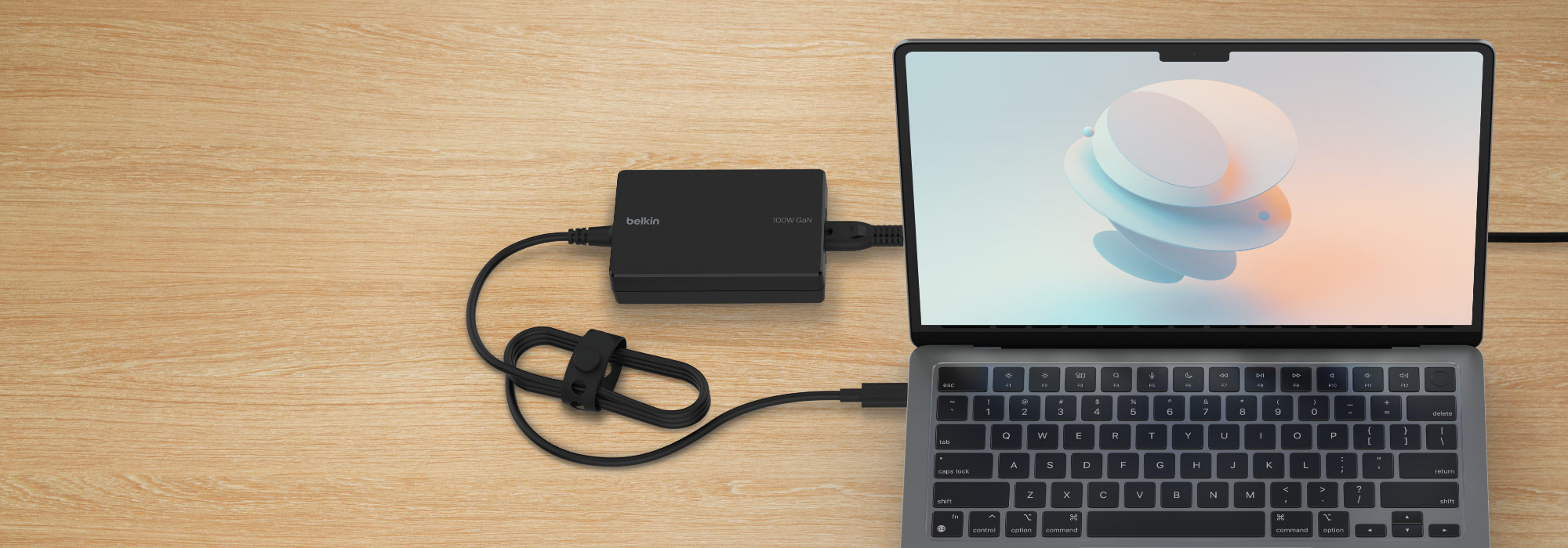 A laptop connected to a Belkin 100W GaN power adapter placed on a wooden surface. The laptop screen displays a pastel abstract 3D shape. The power adapter is plugged into the laptop via USB-C, with the power cord neatly coiled and secured.