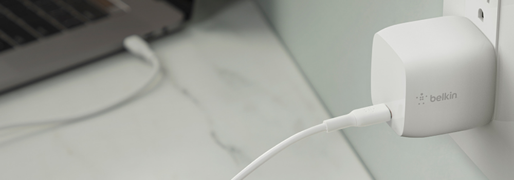 The image displays a white Belkin wall charger connected to an electrical outlet, with a USB-C cable plugged into it. The cable leads toward a laptop in the background, which is partially visible on a clean, marble-textured surface. The design emphasizes simplicity, functionality, and compatibility, with the Belkin charger featuring a sleek and compact form factor. The soft color tones of the setting create a minimalist and modern workspace aesthetic.