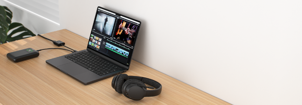 A workspace setup featuring a laptop running video editing software, a pair of black headphones, and a Belkin power bank connected to the laptop via a charging cable. The power bank displays a green battery indicator. The scene is set on a wooden desk with a minimalistic and clean aesthetic.