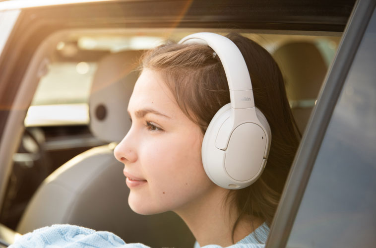 Teen girl wearing white over-ear headphones, looking peacefully out of a car window with sunlight streaming in, enjoying music during a car ride.