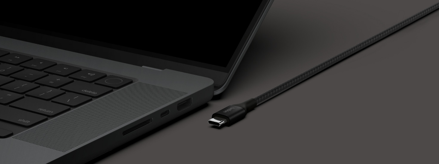 Close-up of a black braided Belkin USB-C cable's connector positioned next to a MacBook Pro's USB-C port, ready to plug in. Dark, moody studio lighting.
