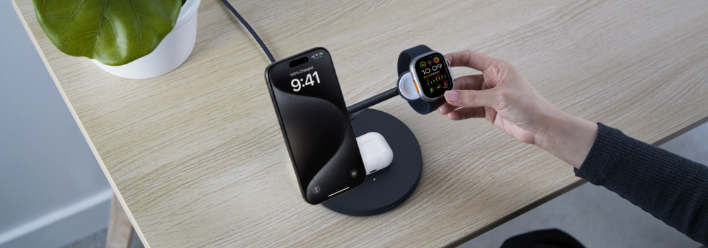This image shows a minimalist charging setup on a wooden desk. A sleek charging dock is holding a smartphone, a pair of wireless earbuds, and a smartwatch simultaneously. The phone displays the time as 9:41, and the smartwatch shows a colorful fitness interface. A hand is seen interacting with the smartwatch. A green plant in a white pot is placed on the desk, adding a natural touch to the modern setup.