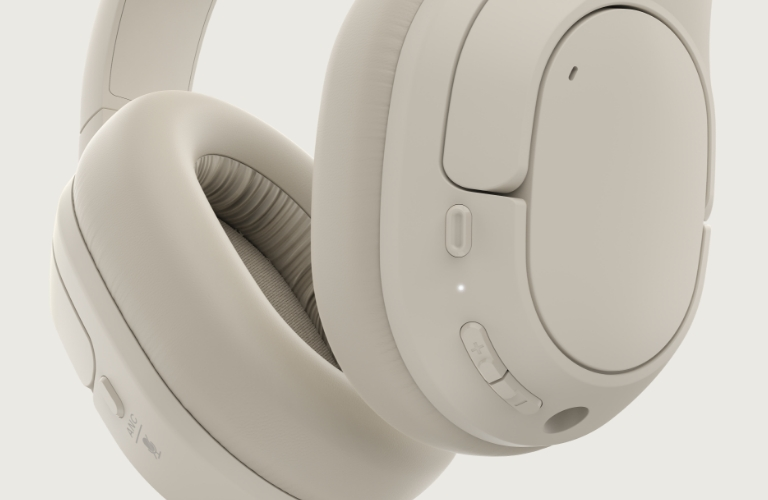 The image shows a close-up view of over-ear headphones in a minimalist beige design. Visible features include soft padded ear cushions, a sleek control panel with buttons for volume adjustment, active noise cancellation (ANC) toggle, and a microphone symbol. The headphones have a modern, ergonomic design with a focus on comfort and functionality.
