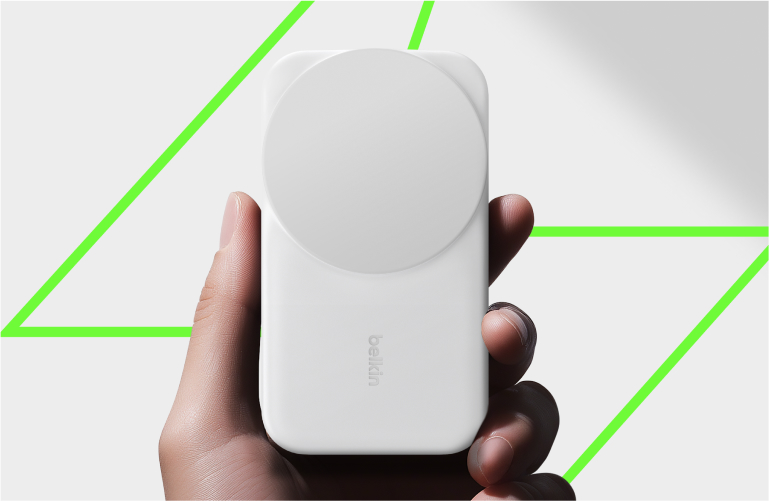 Hand holding a white Belkin magnetic wireless power bank, shown against a bright background with a green geometric outline.