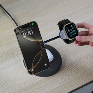 A person places an Apple Watch onto a Belkin 3-in-1 wireless charging stand on a wooden desk. The stand is already charging an iPhone and a pair of AirPods. A potted plant is partially visible in the background.