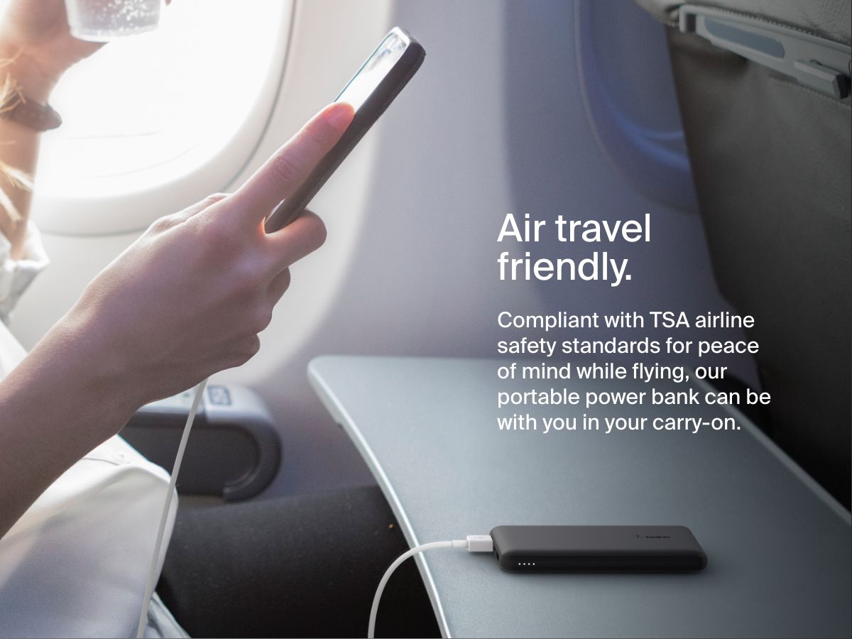 A person uses a smartphone on an airplane while a black Belkin BoostCharge Power Bank 10K rests on the tray table, illustrating its TSA-compliant, travel-friendly design for carry-on use.
