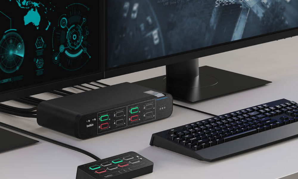 A secure workstation setup featuring a Belkin multiclave KVM switch connected to two monitors, a keyboard, and a remote control pad. The monitors display surveillance and data interfaces, while the KVM switch and control pad show active secure channel indicators with red and green lights for managing multiple network sources.