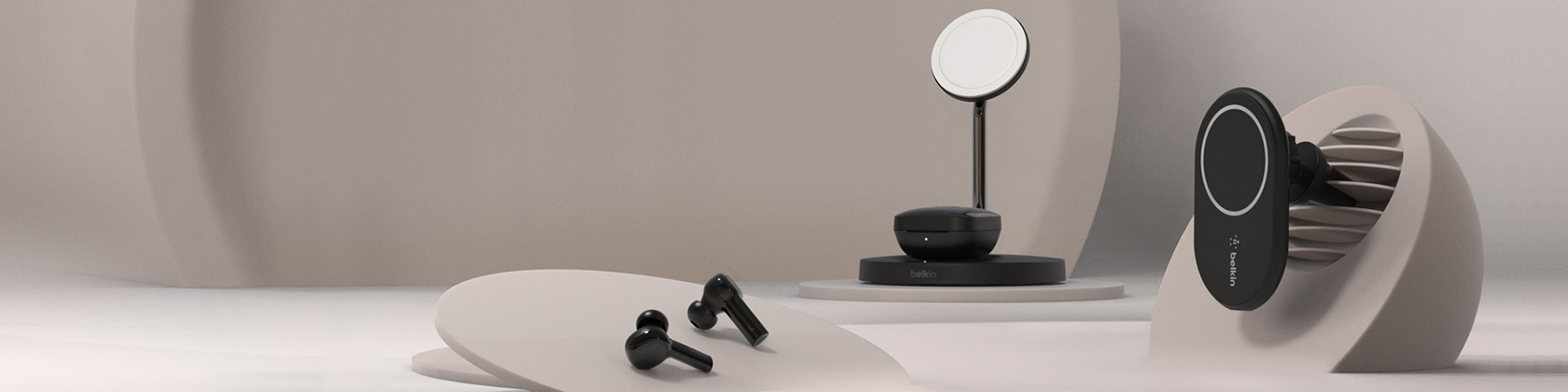 Studio product shot featuring a Belkin BoostCharge Pro MagSafe 2-in-1 wireless charging stand centered on a black pedestal, black wireless earbuds in front, and a separate MagSafe car-vent mount on the right. Beige and grey backdrop with sculptural paper shapes.