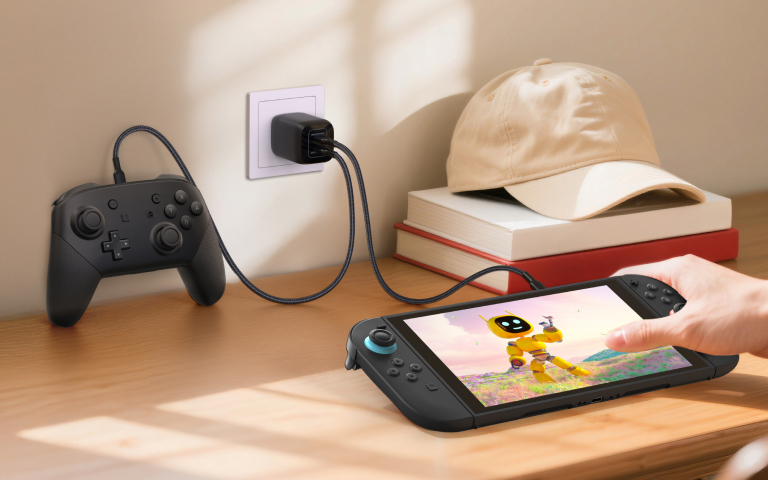 A Nintendo Switch 2 and a game controller are charging on a wooden desk near a sunlit window. The console is being held by a person's hand and displays a bright, cheerful scene featuring a yellow robot in a colorful field. A black USB charging cable connects both devices to a wall adapter plugged into a power outlet. Nearby, a beige baseball cap rests on a stack of books, creating a cozy and organized gaming setup.