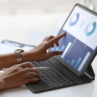 The image features a person using a tablet with a keyboard folio case. The screen displays colorful charts and graphs, indicating data analysis or productivity work. The individual is interacting with the touchscreen, with one hand pointing and the other on the keyboard. The setup is placed on a bright, minimalist desk. A pen is visible in the background, adding to the professional workspace vibe.
