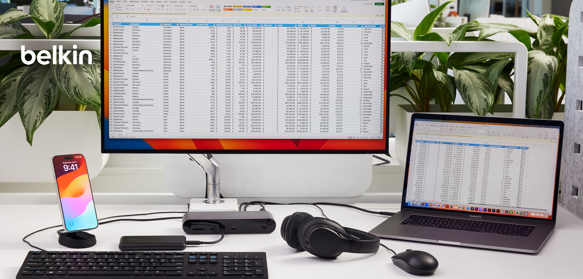 A modern office desk setup featuring a large external monitor displaying an Excel spreadsheet, a MacBook laptop also showing a spreadsheet, and various accessories. The workspace includes a Belkin docking station, a wireless keyboard, a mouse, a pair of headphones, and a smartphone on a charging stand. The environment is well-lit, with green plants in the background, creating a clean and organized workspace.
