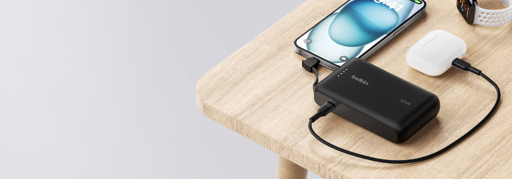 A black Belkin 20W power bank placed on a wooden table, charging a smartphone and a pair of wireless earbuds via USB-C cables. A smartwatch with a white band is partially visible in the background. The scene highlights the compact and portable design of the power bank.