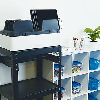 This image showcases a modern classroom setup featuring a Belkin device charging station holding several tablets. The charging station is neatly placed on a black stand, positioned beside a white cubby storage unit. The background includes plants, containers, and organized school supplies, reflecting an efficient and clean educational environment. The Belkin logo is prominently displayed in the upper-left corner.