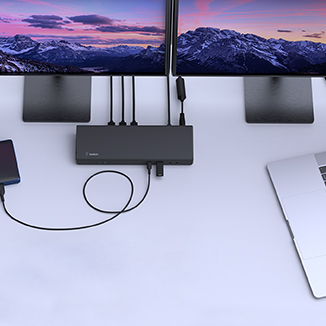 A Belkin docking station set up on a modern workspace, connecting a MacBook to dual monitors displaying a scenic mountain landscape. The docking station also links to an external hard drive and multiple accessories, showcasing its multi-device connectivity and expanded workstation capabilities.