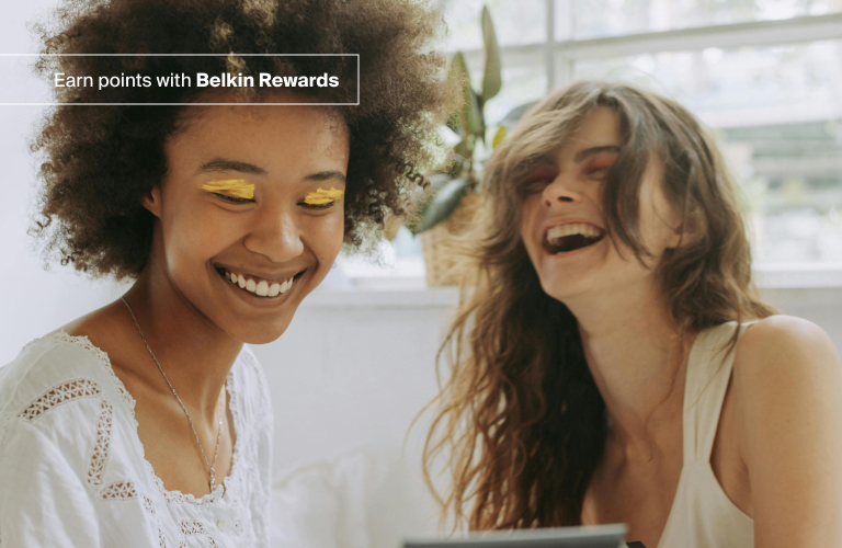 Two women laughing together indoors, one with curly hair and bright yellow eyeshadow, while a promotional text overlay reads, "Earn points with Belkin Rewards."