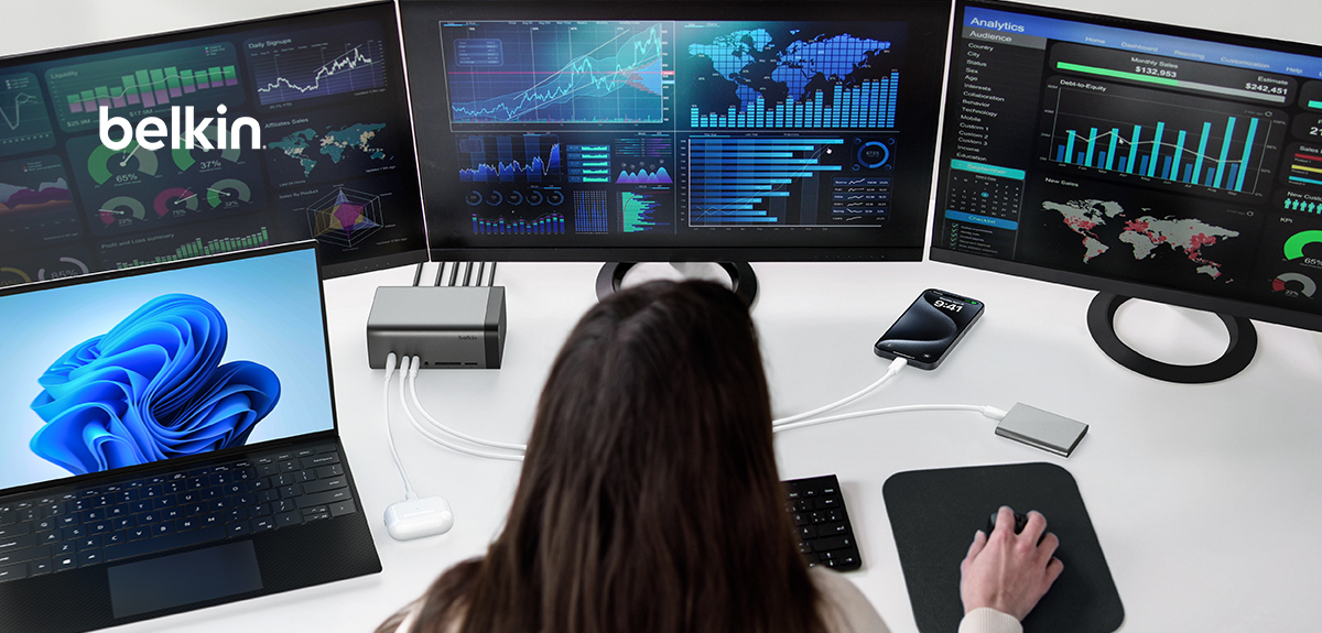 A person using a multi-monitor setup connected to a Belkin docking station. The setup includes a laptop, three monitors displaying various data analytics and charts, and connected devices such as a smartphone and wireless earbuds. The person is interacting with the setup using a mouse and keyboard, creating an efficient and organized workspace for data analysis or trading.