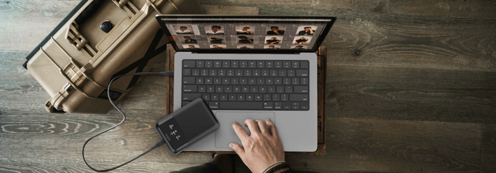 This image shows a laptop being used on a wooden surface, with a hand operating the trackpad. The laptop is connected to an external hard drive via a USB cable. The screen displays thumbnails of photos, suggesting the user is working with media files. Next to the laptop is a sturdy, beige protective case, possibly for equipment or gear.