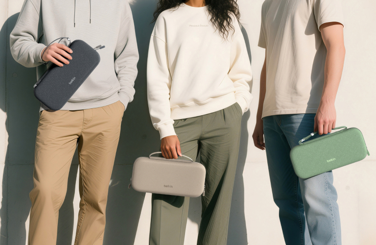 Three individuals stand against a sunlit, minimalist concrete wall, each holding a Belkin protective carrying case for the Nintendo Switch 2 in different colors—dark gray, light beige, and green. The image emphasizes modern, on-the-go protection and style for portable gaming.