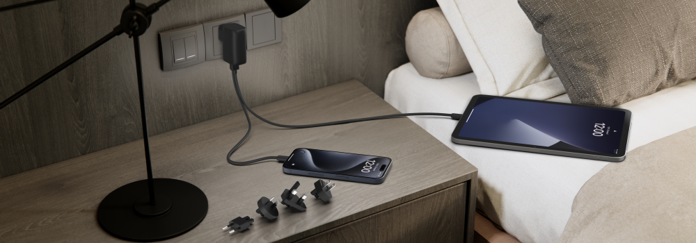 A nightstand setup featuring a Belkin wall charger plugged into an outlet, charging both a smartphone and a tablet via USB-C cables. Multiple plug adapters are placed on the table, indicating international compatibility. A modern black lamp and a neatly arranged bed are visible in the background, creating a cozy and functional bedside charging station.