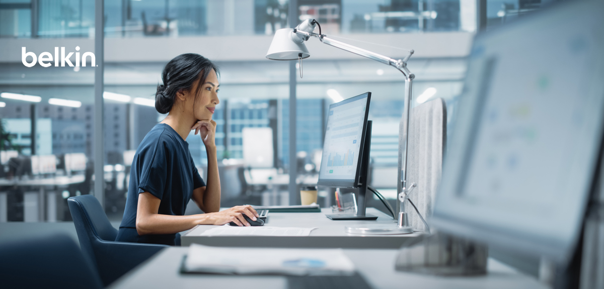 Person working at a modern office desk using a desktop computer, viewed in profile, with the Belkin logo displayed in the upper left and a bright, open workspace in the background.