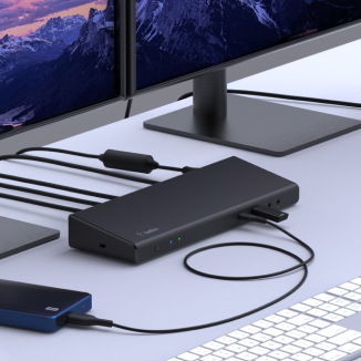 The image showcases a sleek docking station connected to multiple devices, including monitors, a laptop, and an external storage device. The setup is placed on a clean desk with a white keyboard in the foreground, emphasizing a modern and organized workspace. The background includes dual monitors displaying a scenic mountain view.