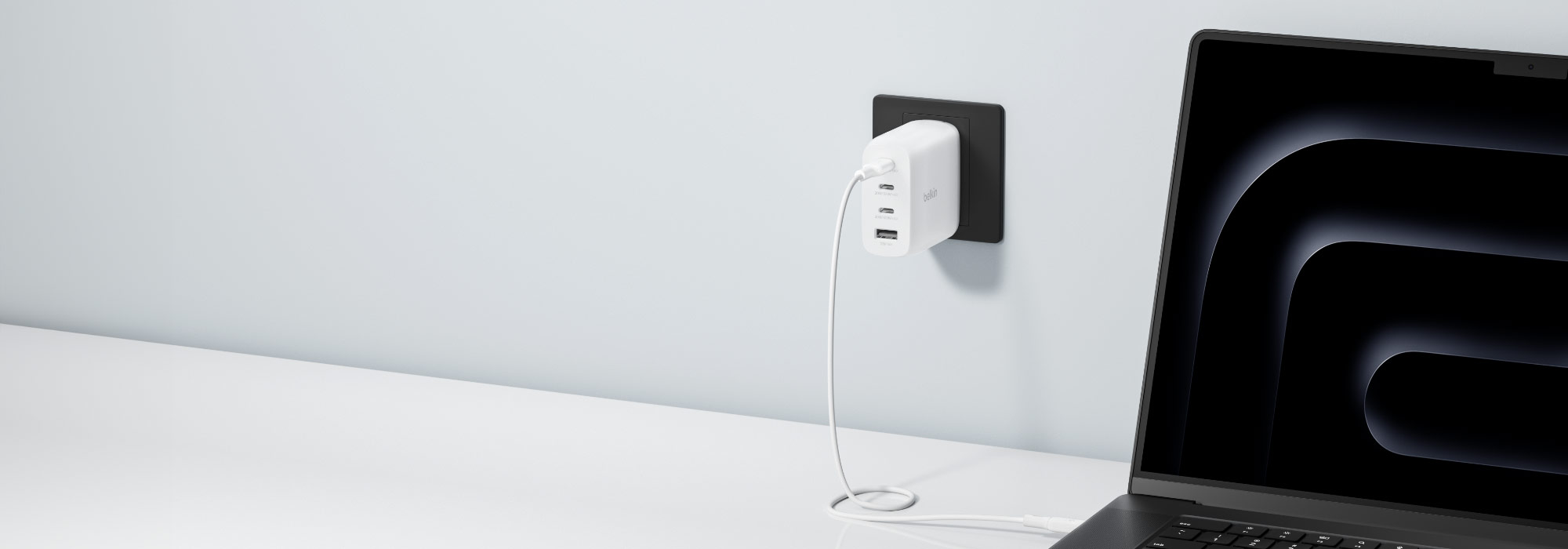 A laptop connected to a white Belkin wall charger plugged into a black power outlet on a light-colored wall. The charger has multiple ports and a white cable connecting it to the laptop, which displays a dark abstract design on its screen. The setup rests on a clean white desk.