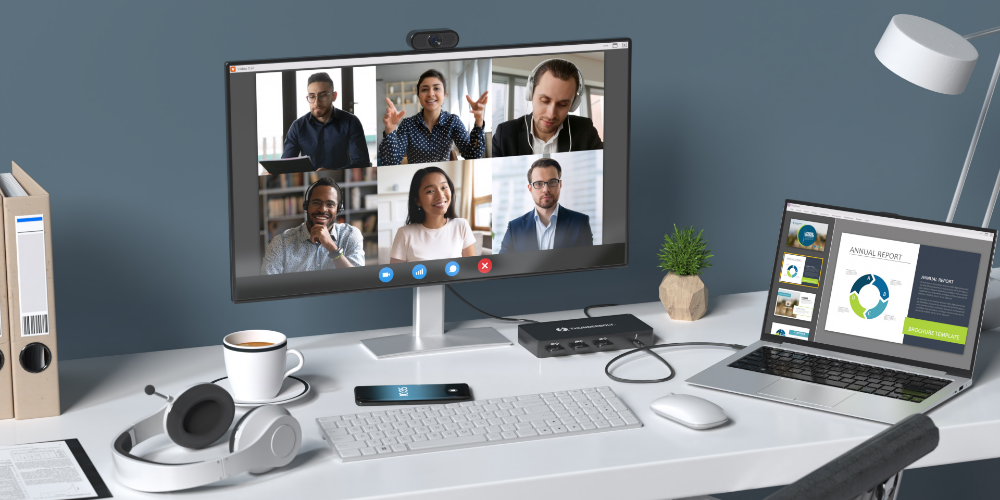 A modern home office setup designed for remote work and video conferencing. The desk features a large monitor displaying a virtual meeting with six participants, a laptop open to an annual report presentation, a wireless keyboard and mouse, and a docking station. A pair of white headphones rests on the desk next to a cup of coffee and a smartphone. The workspace is neatly organized with a minimalist aesthetic, including a small plant, file folders, and a sleek desk lamp against a dark blue wall.