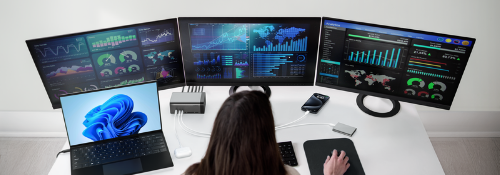 Person working at a desk with a laptop and three large monitors displaying data dashboards, connected through a docking station with multiple devices including a smartphone.