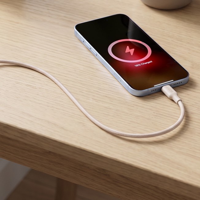 A white Belkin compact USB-C wall charger plugged into a wall outlet, its braided cable running across a light oak desk to an iPhone lying face up with its charging screen on, next to a small plant, a mug and a notebook