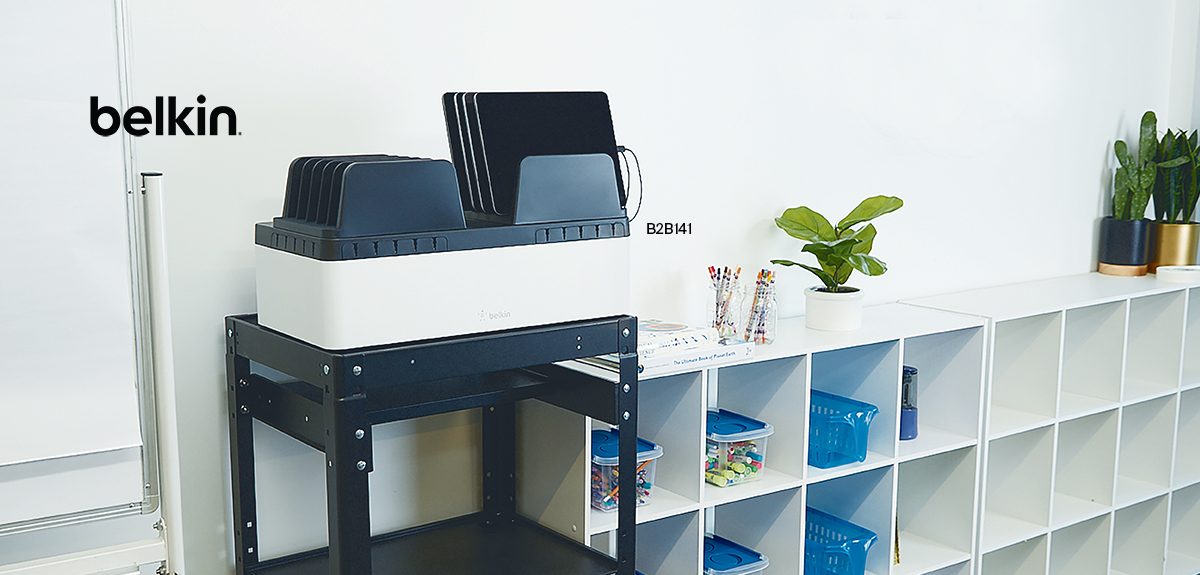 This image showcases a modern classroom setup featuring a Belkin device charging station holding several tablets. The charging station is neatly placed on a black stand, positioned beside a white cubby storage unit. The background includes plants, containers, and organized school supplies, reflecting an efficient and clean educational environment. The Belkin logo is prominently displayed in the upper-left corner.