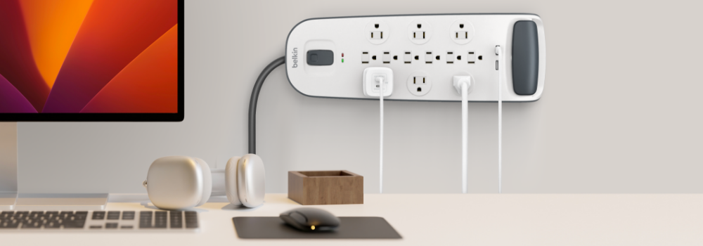 A modern workspace featuring a wall-mounted Belkin surge protector with multiple outlets and USB ports, plugged in with several white charging cables. On the desk below are a computer monitor, wireless headphones, a wooden accessory holder, a wireless mouse on a pad, and part of a keyboard. The background is minimalistic with neutral tones.
