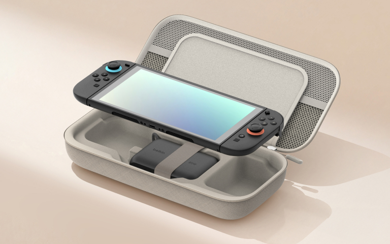 A handheld gaming console with attached controllers is neatly placed inside a light gray, open carrying case. The console screen is reflective with a soft gradient, and the controllers feature colorful joystick rings (blue and orange). A compact black charging accessory labeled "Belkin" is secured inside the case with an elastic strap. The scene is set against a minimal, softly lit beige background.